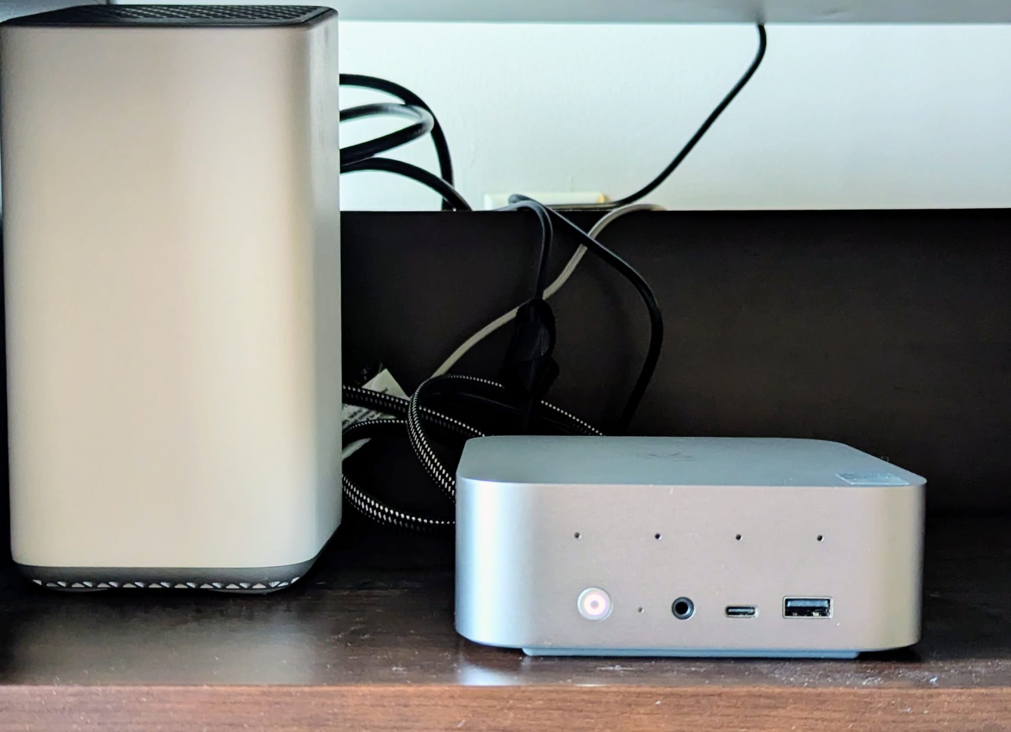 A small silver Beelink mini PC sitting on a dark wooden shelf beside a tall white home router, with cables coiled behind them. The mini PC is roughly a third the height of the router.